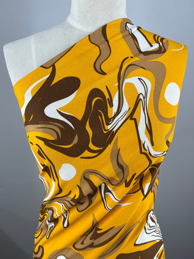 A mannequin is adorned with the Printed Crepe - Cappuccino fabric from Super Cheap Fabrics, showcasing a vibrant abstract design with swirling patterns in shades of orange, brown, white, and yellow. The gray background enhances the bold colors of the 150 cm polyester crepe fabric.