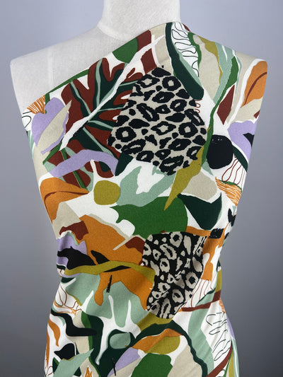 A mannequin is adorned in a one-shoulder garment made from Super Cheap Fabrics' "Printed Crepe - Splash - 150cm," featuring a striking, multicolored pattern. The design includes leopard spots, abstract shapes, and a mix of green, orange, purple, and white hues, creating a vibrant and artistic high fashion look.