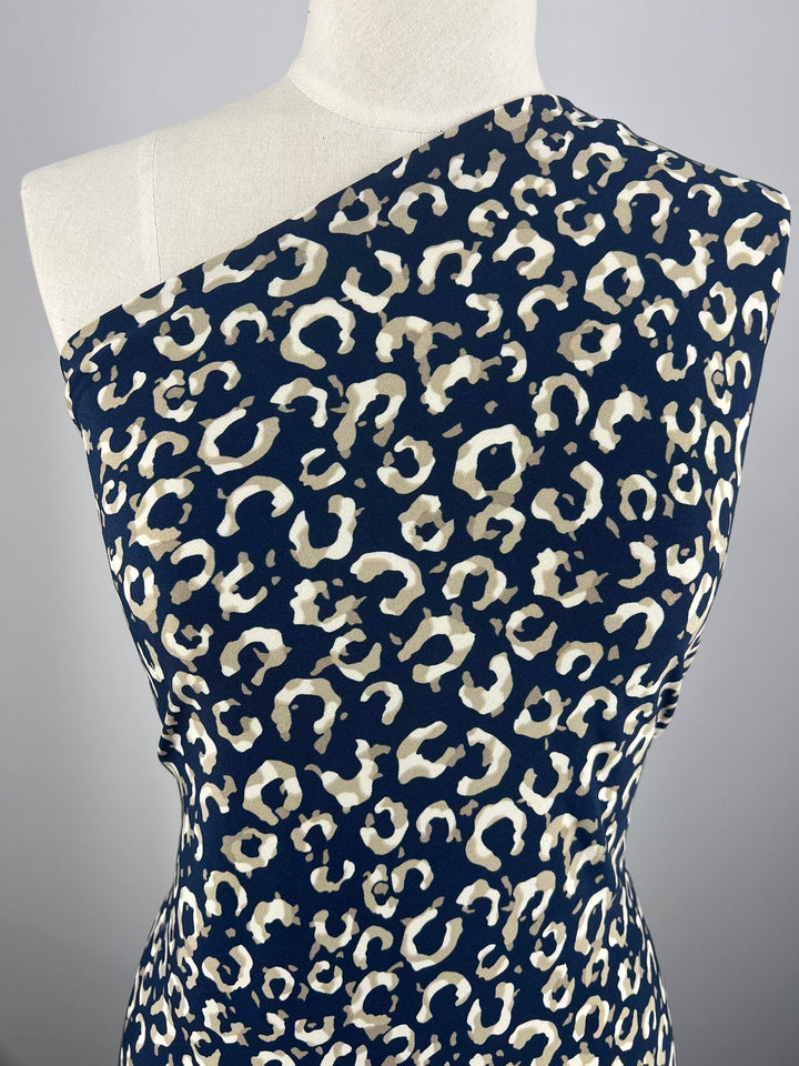 A mannequin wearing a Printed Lycra - Jungle - 150cm crafted from medium-weight Polyester/Spandex fabric with a leopard print design in shades of white and beige on a dark blue backdrop. The print features irregular spots, creating a bold, fashionable look against the plain gray background. This exquisite fabric is offered by Super Cheap Fabrics.