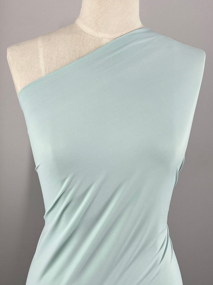 A mannequin draped in a Clearwater-colored, one-shoulder dress made from Super Cheap Fabrics' Nylon Lycra - 155cm is positioned against a plain, light gray background. The fabric has a slight sheen and smooth texture, elegantly wrapping around the torso with four-way stretch for added comfort and flexibility.