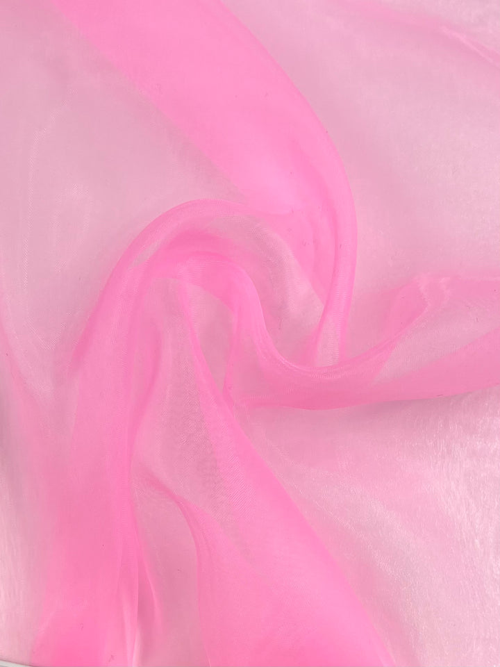 A close-up of the Super Cheap Fabrics' Organza - Sachet Pink - 150cm shows its translucent fabric with soft, flowing folds. The lightweight and delicate sheer organza material creates gentle curves and a subtle sheen in the light.