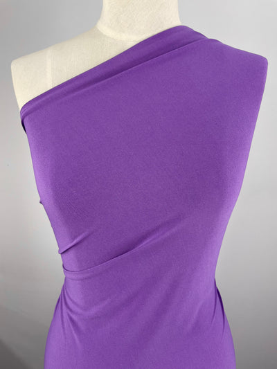A mannequin draped in a vibrant Super Cheap Fabrics' ITY Knit - Royal Lilac - 150cm, one-shoulder, medium-weight fabric. The fabric is smoothly wrapped around the upper torso, creating elegant folds and highlighting the body's natural shape. The background is plain and neutral, emphasizing the garment's rich color.